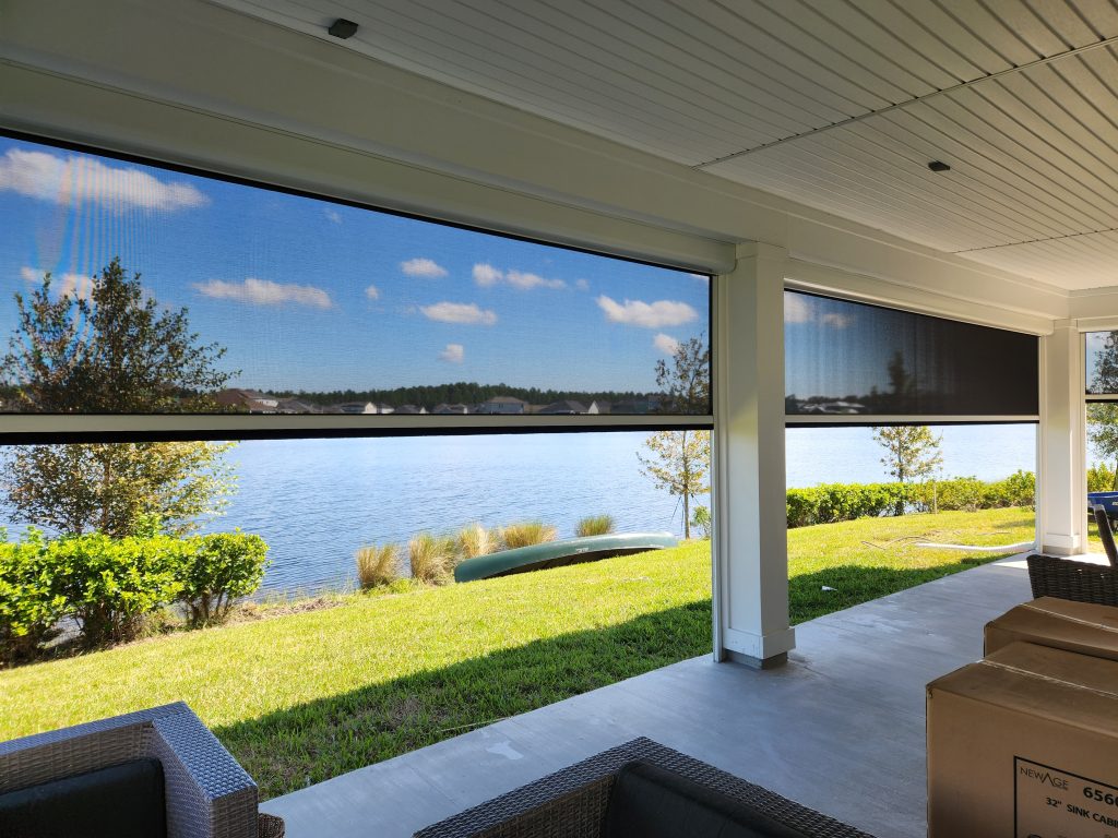 A covered patio with wicker furniture overlooks a lake, framed by large retractable screens. Green grass, shrubs, and trees line the shore, and blue sky with scattered clouds is visible in the background.