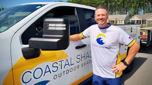 Todd Mason in a white and yellow Coastal Shades Outdoor Shades shirt stands beside a matching company truck on a sunny day, with houses and trees visible in the background.